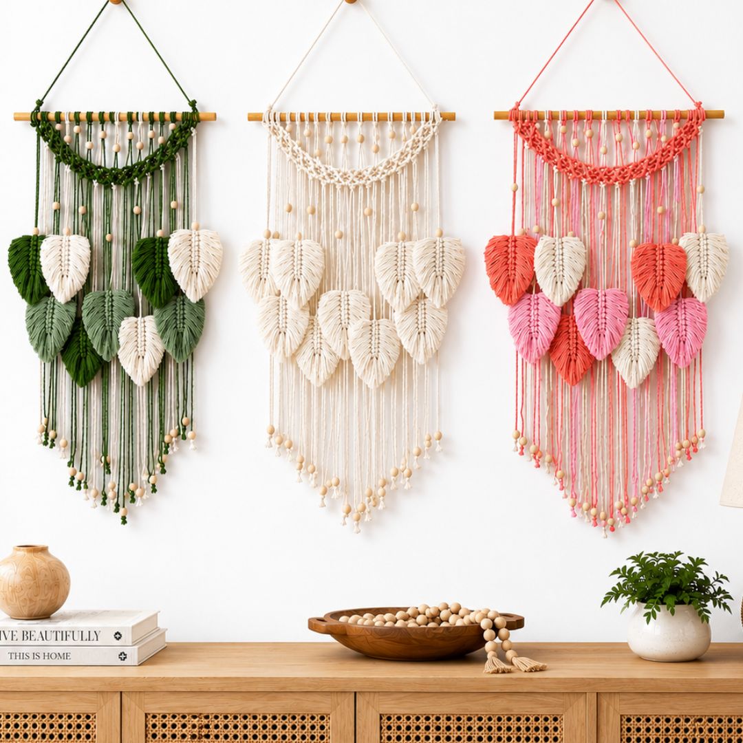 Three woven wall hangings with leaf-shaped macrame details are displayed on a white wall above a wooden cabinet. The left one is green and white, the middle is white, and the right one is peach, white, and pink.