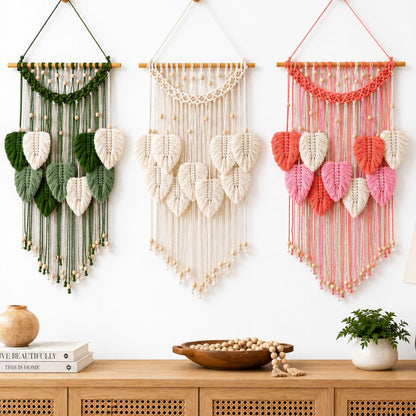Three woven wall hangings with leaf-shaped macrame details are displayed on a white wall above a wooden cabinet. The left one is green and white, the middle is white, and the right one is peach, white, and pink.