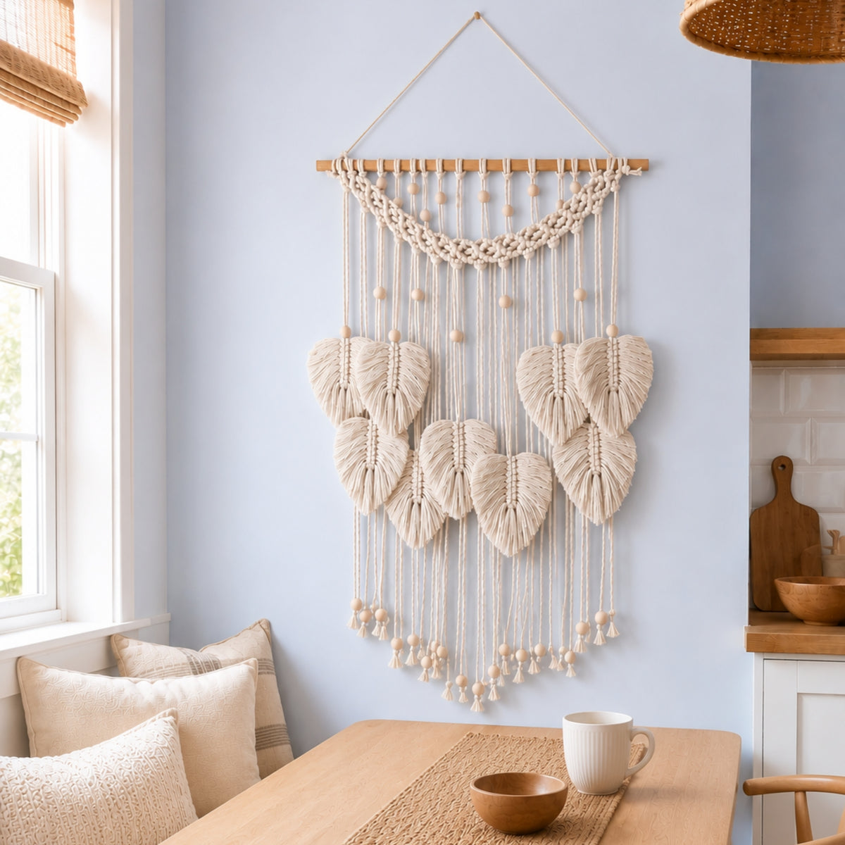 A bohemian-style white macrame wall hanging with knotted leaf shapes and wooden beads, suspended from a wooden rod in a bright, modern room with a light blue wall, wooden table, and neutral cushions.